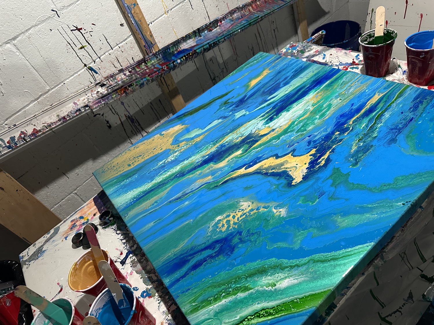A poured painting in blues, greens, and gold resting on a paint-covered studio table, surrounded by cups of paint.