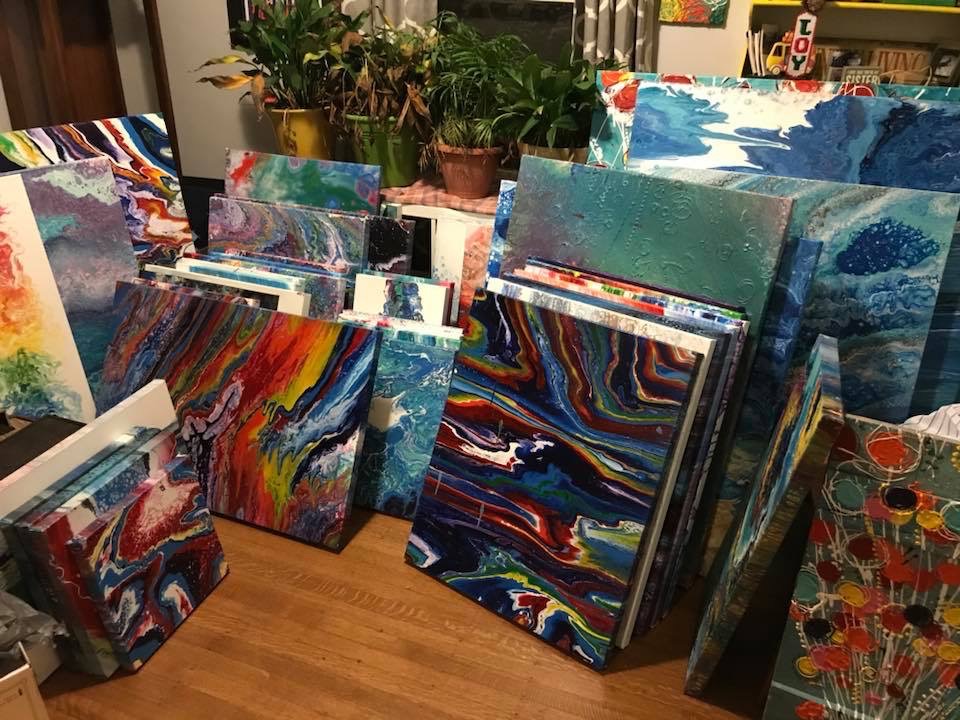 Many finished paintings leaning against one another on a studio floor, in bright poured colours.